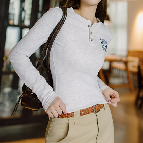 Person wearing a light gray long-sleeve shirt with a logo, beige pants, and carrying a brown bag indoors.